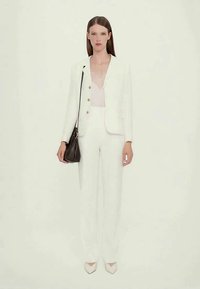 Femme portant un costume blanc ajusté avec une blouse transparente en dessous, tenant un sac à main noir, se tenant devant un fond uni.