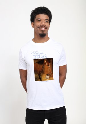 Mężczyzna w białym t-shircie z nadrukiem obrazu Edgara Degasa „W Stajniach” przedstawiający konia i psa w stajni.