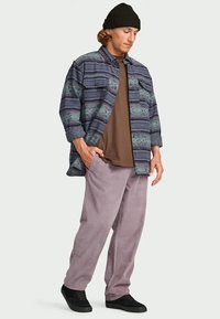 Homem a usar um gorro preto, uma camisa de botão riscada sobre uma t-shirt castanha, calças lavanda e sapatos pretos, de pé com uma mão no bolso.