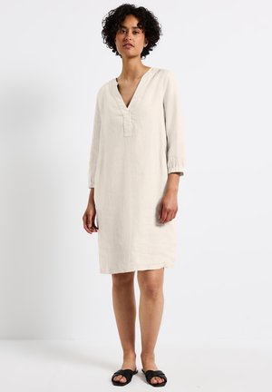 Beige linneklänning med trekvartslånga ärmar, V-halsringning och lös passform. Klänningen slutar ovanför knäet och har en subtil textur. Bärs med svarta sandaler.