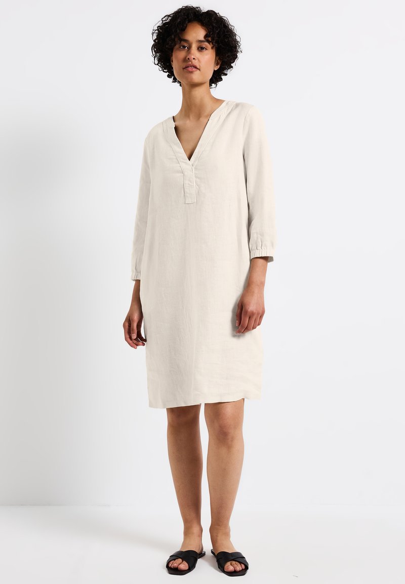 Beiges Leinenkleid mit 3/4-Ärmeln, V-Ausschnitt und lockerer Passform. Endet oberhalb des Knies und weist eine dezente Textur auf. Getragen mit schwarzen Pantoletten.