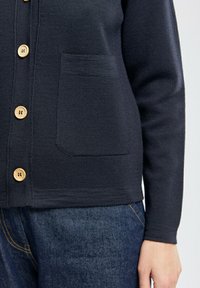 Gilet en tricot bleu marine avec des boutons en bois et une poche avant, porté sur un jean en denim bleu foncé, montrant la manche et le côté du torse.