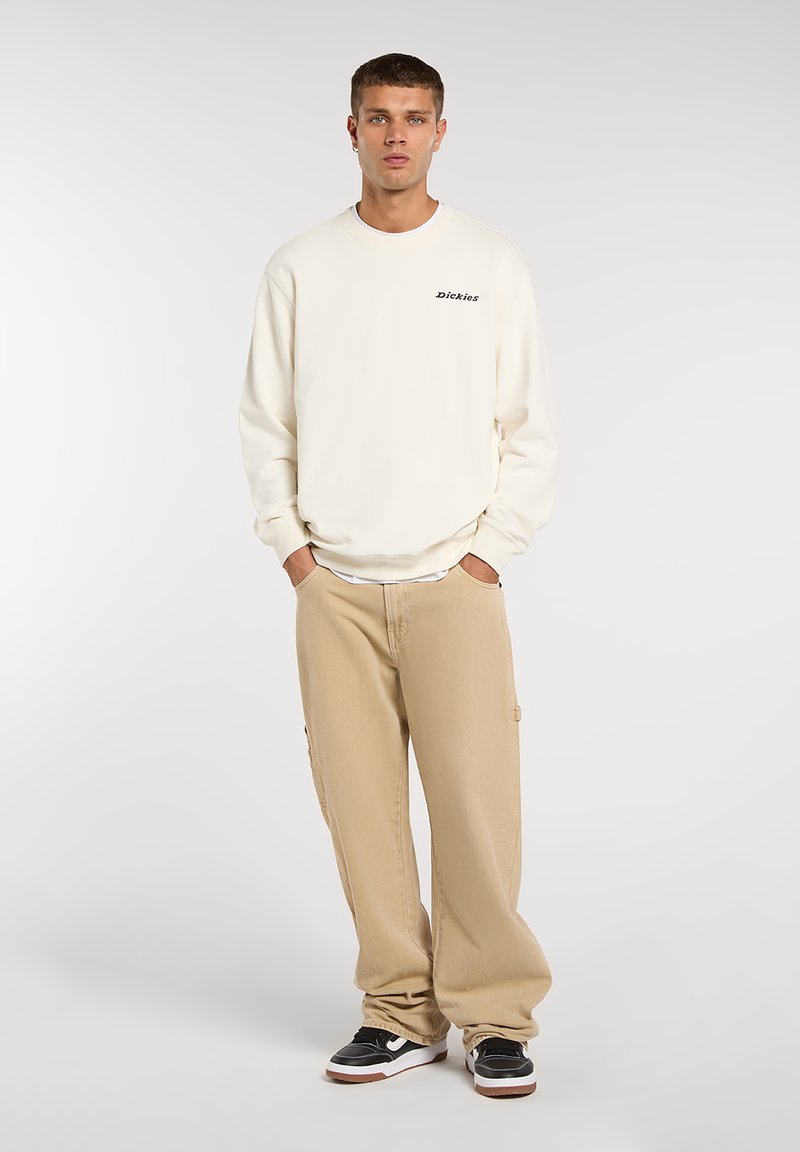 Sudadera color crema con el logo de "Dickies", combinada con pantalones cargo beige sueltos y zapatillas negras con detalles blancos. Posando de pie contra un fondo claro.