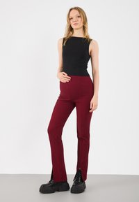 Black sleeveless top and burgundy ribbed pants with side slits. Pants feature a fitted design and are paired with chunky black footwear.