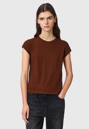 Junge Frau mit glatten dunklen Haaren, die ein braunes kurzärmeliges Shirt und dunkle Jeans trägt, mit einer Hand in der Tasche, stehend vor hellem Hintergrund.
