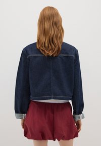 Veste en denim foncé, cropped, avec des manches longues, des poignets à bouton et des coutures contrastées. Assortie à un short rouge ample.
