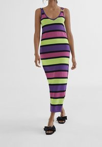 Kvinna iklädd en ärmlös V-ringad klänning i ankellängd med horisontella lila, gröna, rosa och svarta ränder, samt svarta rosettsandaler.