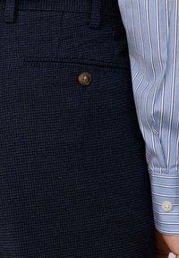 Tascone blu navy testurizzato con un bottone marrone e una manica di camicia a righe blu e bianche con un polsino bianco con bottone.