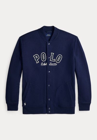 Μπουφάν σε ναυτικό μπλε χρώμα με κουμπιά και κείμενο "POLO Ralph Lauren" στο στήθος, δύο μπροστινές τσέπες, ριμπ κολάρο και μανσέτες, και μικρό λογότυπο κοντά στο τελείωμα.