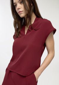 Top corto bordeaux con scollo a V, realizzato in un tessuto morbido. Presenta una vestibilità comoda e un orlo discreto. Il modello ha le mani nelle tasche.