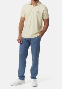 Hellgelbes Poloshirt mit Kragen und dezentem Streifenmuster, kombiniert mit blauen Nadelstreifenhosen und weißen Turnschuhen.