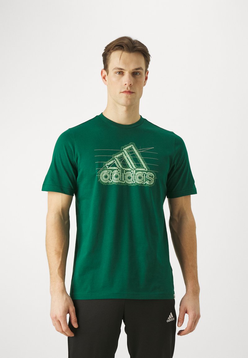 Camiseta de algodón verde con mangas cortas, que presenta un destacado logo de Adidas en blanco contorneado a lo largo del pecho. Cuello redondo, ajuste holgado.