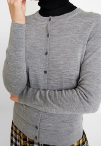 Grå cardigan gjord av mjukt stickat tyg med rund halsringning, knappad front och långa ärmar, med subtila ribbade muddar och nederkant.