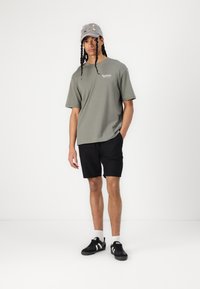 Olivengrøn kortærmet t-shirt, sorte shorts, sorte sko med hvide striber og en grå kasket med farverig broderi.