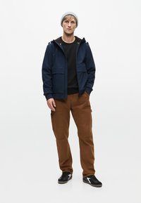 Marineblå zip-jakke med to frontlommer, sort crewneck t-shirt, brune cargo-bukser og sorte sneakers med hvide detaljer. Grå beanie.