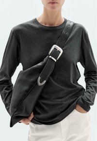 Schwarze Leder-Schultertasche mit mattierter Oberfläche, großem silbernen Schnallenverschluss und konischer Form, getragen über einem dunklen Langarmoberteil.