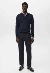 Man staat in een marineblauwe pullover over een lichtblauw shirt, donkere broek en zwarte instappers tegen een witte achtergrond.