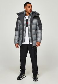 Grijze pufferjack met een glanzende afwerking, voorzien van een zwarte capuchon en schouderaccenten; gedragen met zwarte cargobroeken en sportieve schoenen.