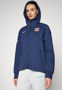 Navy zip-up hoodie with a high collar, featuring a Barcelona logo and orange accents. It has a front zipper and side pockets.