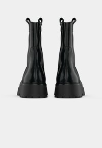 Schwarze Lackleder-Stiefeletten mit einer massiven Sohle, Zuglaschen und einem ausgeprägten Absatz. Verfügt über ein elegantes, glattes Finish und genähte Details.