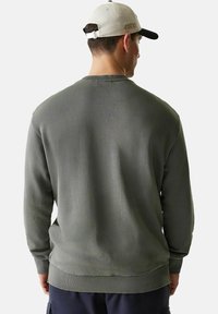 Mann trägt einen olivgrünen Sweatshirt und eine hellgraue und schwarze Baseballkappe, von hinten gesehen vor einem einfarbigen Hintergrund.