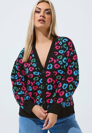 Vrouw die een zwarte cardigan draagt met een kleurrijke roze, blauwe en groene luipaardprint en drie knopen, gecombineerd met een blauwe spijkerbroek.