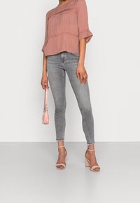 Blouse texturée rose clair avec des manches à volants, associée à un jean skinny gris. Tenant un petit sac à main rose et portant des sandales à talons blocs rose pâle.