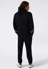 Felpa nera con tasca frontale e dettagli grigi sulle maniche, abbinata a pantaloni jogger neri e sneaker bianche, vista da dietro.