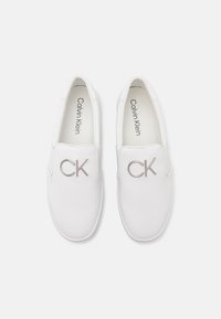 Hvide slip-on sko lavet af glat materiale, med en sølvfarvet "CK"-logo foran og rund tå-design.