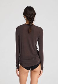 Cămașă maro cu mâneci lungi dintr-un material moale și ușor, cu croială lejeră și design fără cusături, având un decolteu rotunjit și o parte din spate prelungită.