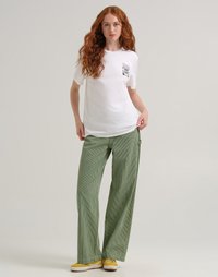 Giovane donna con lunghi capelli rossi, che indossa una maglietta bianca con grafica, pantaloni a gamba larga a righe verdi e sneaker gialle, in piedi davanti a uno sfondo semplice.