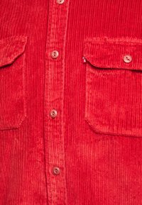 Camisa de pana roja con botones en la parte delantera, dos bolsillos en el pecho y amplias líneas verticales. Presenta botones redondos y una textura suave.