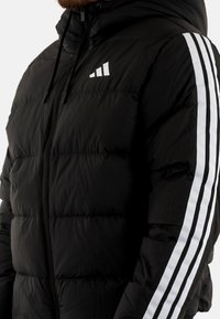 Chaqueta acolchada negra con cierre de cremallera, diseño acolchado y capucha. Presenta detalles de rayas blancas en las mangas y un logo en el pecho.