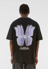 Sort bomulds t-shirt med et stort sommerfuglegrafik i lys lilla på ryggen, med hvid tekst underneath. Korte ærmer og afslappet pasform.