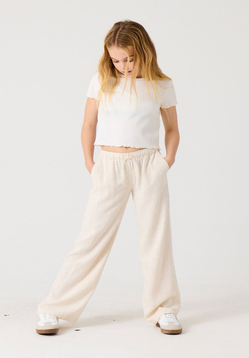 Fille aux longs cheveux blonds regardant vers le bas, portant un crop top blanc à manches courtes, un pantalon ample beige et des baskets blanches, debout les mains dans les poches.