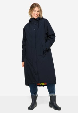 Navy long coat with a hood, zip front, and drawstring. Features side pockets and a vibrant inner lining. Paired with denim and black rubber boots.