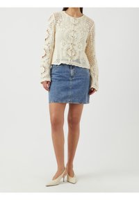 Szydełkowy kremowy sweter z długimi rękawami i zapięciem na guziki, zestawiony z jasnoniebieską dżinsową spódnicą mini i beżowymi butami na obcasie.