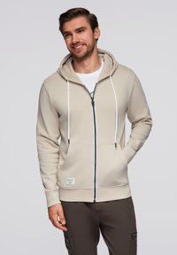 Felpa beige con zip e cappuccio regolabile, dettagli in corda bianca e doppie tasche frontali. Realizzata in un tessuto liscio e morbido.