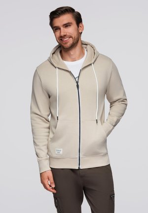 Sudadera con cremallera beige, capucha con cordón, detalles en cordón blanco y doble bolsillo frontal. Fabricada con un tejido suave y liso.
