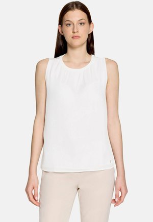 Jonge vrouw met steil bruin haar, die een mouwloze witte blouse en beige broek draagt, staand tegen een effen lichte achtergrond.