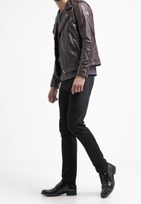 Giacca da motociclista in pelle marrone con cerniere argentate, indossata sopra una maglietta grigia e abbinata a jeans neri slim-fit e scarpe eleganti nere.
