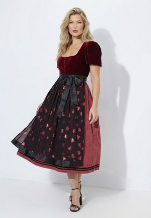 Mujer con un vestido de terciopelo burdeos con mangas abullonadas y falda delantal negra adornada con patrones de corazones rojos, posando sobre un fondo blanco.