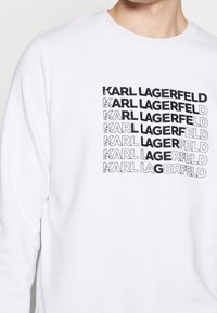 Sudadera blanca de manga larga con el texto negro apilado "KARL LAGERFELD" en diferentes grosores. Tejido suave, cuello redondo.