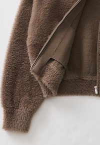 Bruine nepbontjas met een ritssluiting aan de voorkant, met een zachte, gestructureerde buitenkant en gebreide manchetten. Voorzien van een gladde binnenvoering.