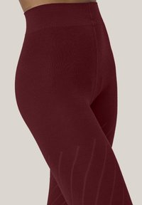 Närbild av en person med hög midja, texturerade, mörkröda leggings mot en enfärgad ljus bakgrund.