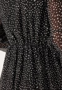 Robe en tissu noir avec une taille froncée, ornée d'un motif à pois de tailles variées et de manches opaques. Texture lisse.