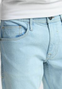 Jeans de mezclilla de color azul claro con una textura suave, un bolsillo para monedas y costuras en contraste. El ajuste parece relajado en la cintura.