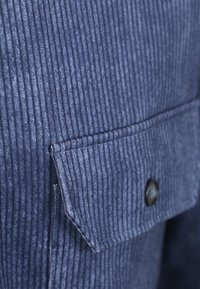 Navyblauwe ribfluweel stof heeft verticale ribbels, met een opgestikte zak en een zwarte knop aan de zijkant. De textuur is zacht en iets geribbeld.