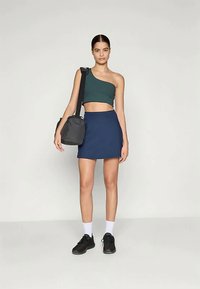 Egy vállra akasztott zöld crop top, amelyet egy tengerészkék mini szoknyával párosítottak. A modellen fekete tornacipő és fekete táska van. Semleges háttér.