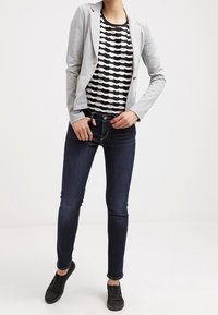 Blazer ajustado gris sobre una blusa de rayas en blanco y negro con patrón ondulado, combinado con jeans de mezclilla oscuros y zapatillas negras.